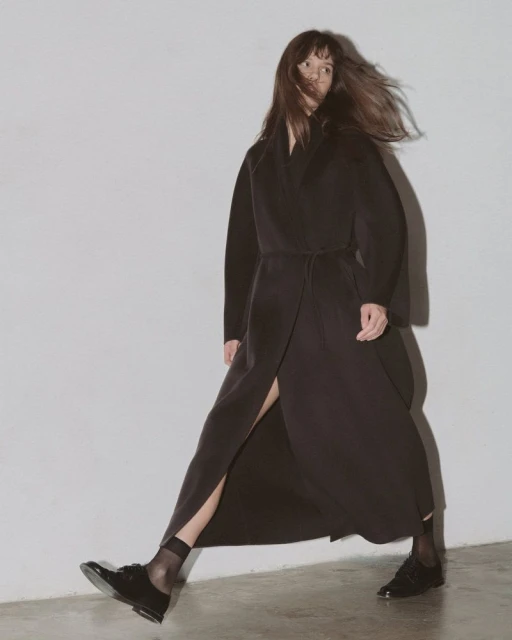 Woman in a long dark coat walks against a white wall, showcasing fashion and style with her dark dress and patent leather shoes.
