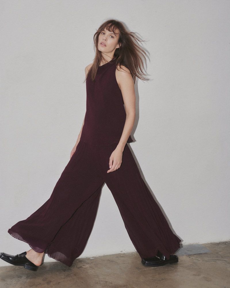 A young woman in a maroon sleeveless top and wide-leg pants walks against a white wall in an indoor studio setting.