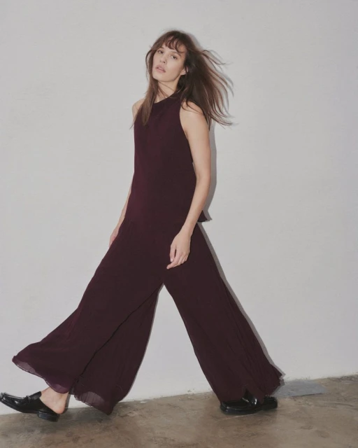 A young woman in a maroon sleeveless top and wide-leg pants walks against a white wall in an indoor studio setting.