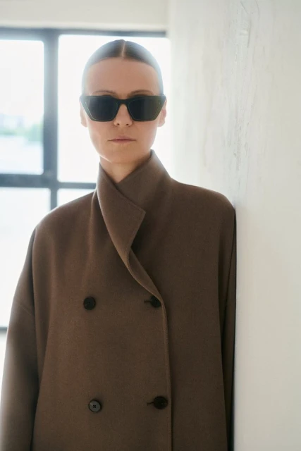 Woman in brown coat and sunglasses stands in neutral indoor setting, leaning against wall, with soft natural window light.