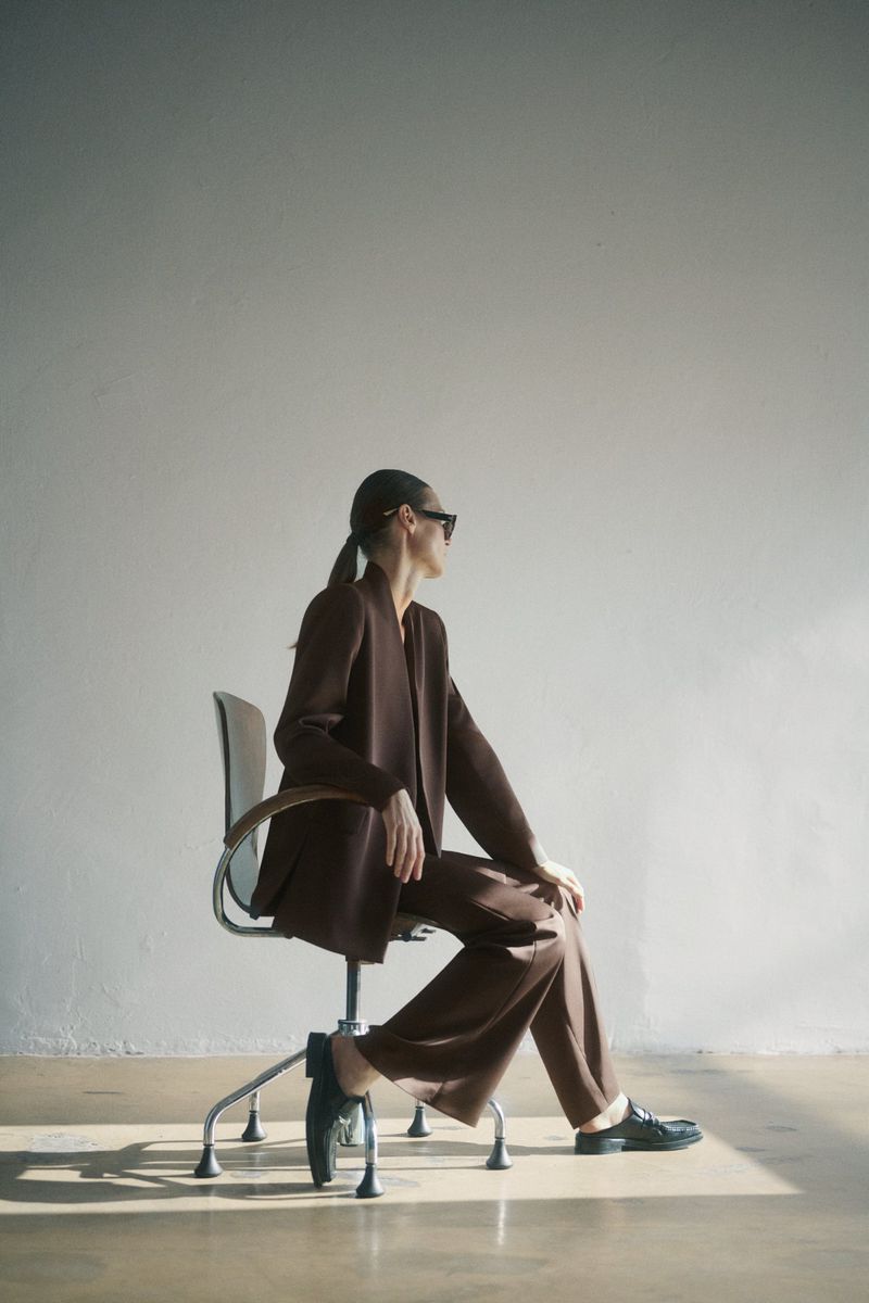 A woman in a brown pantsuit sits on a chair in a sunlit studio. She wears sunglasses and dark flats in this full, brightly lit shot.