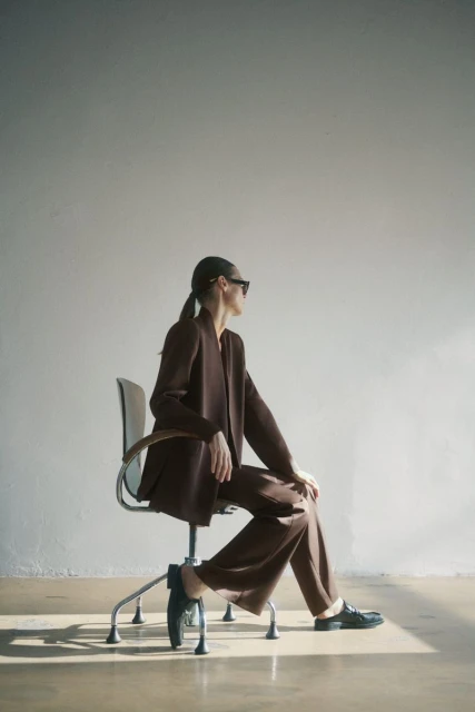 A woman in a brown pantsuit sits on a chair in a sunlit studio. She wears sunglasses and dark flats in this full, brightly lit shot.