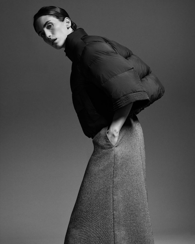 Monochrome fashion shot of a woman wearing a puffy jacket and flared skirt, standing in a photo studio with a simple grey background.