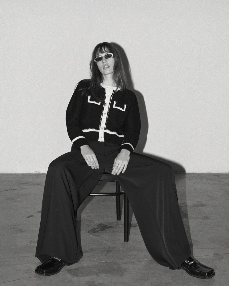Black-and-white portrait of a woman with bangs wearing a black cardigan with white trim, black pants, loafers, and sunglasses, seated on a stool.