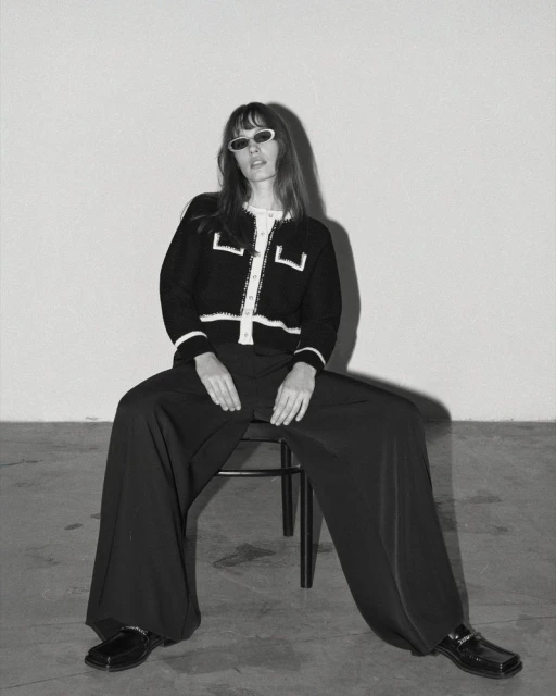 Black-and-white portrait of a woman with bangs wearing a black cardigan with white trim, black pants, loafers, and sunglasses, seated on a stool.