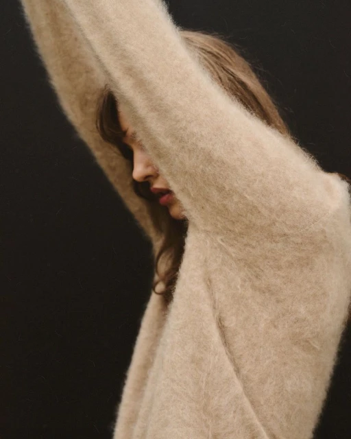 Side profile portrait of a woman in a fuzzy beige sweater raising her arms up in front of a dark background in a fashion style photo.