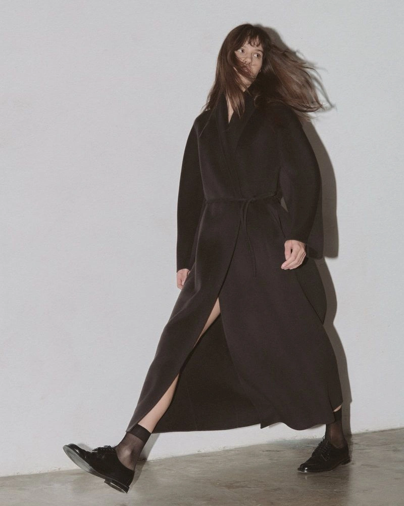 Model wearing a long belted black coat, sheer black socks, and black oxfords striding against a plain studio wall.