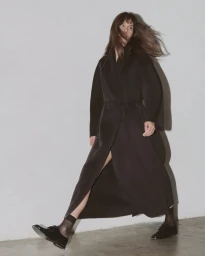 Model wearing a long belted black coat, sheer black socks, and black oxfords striding against a plain studio wall.