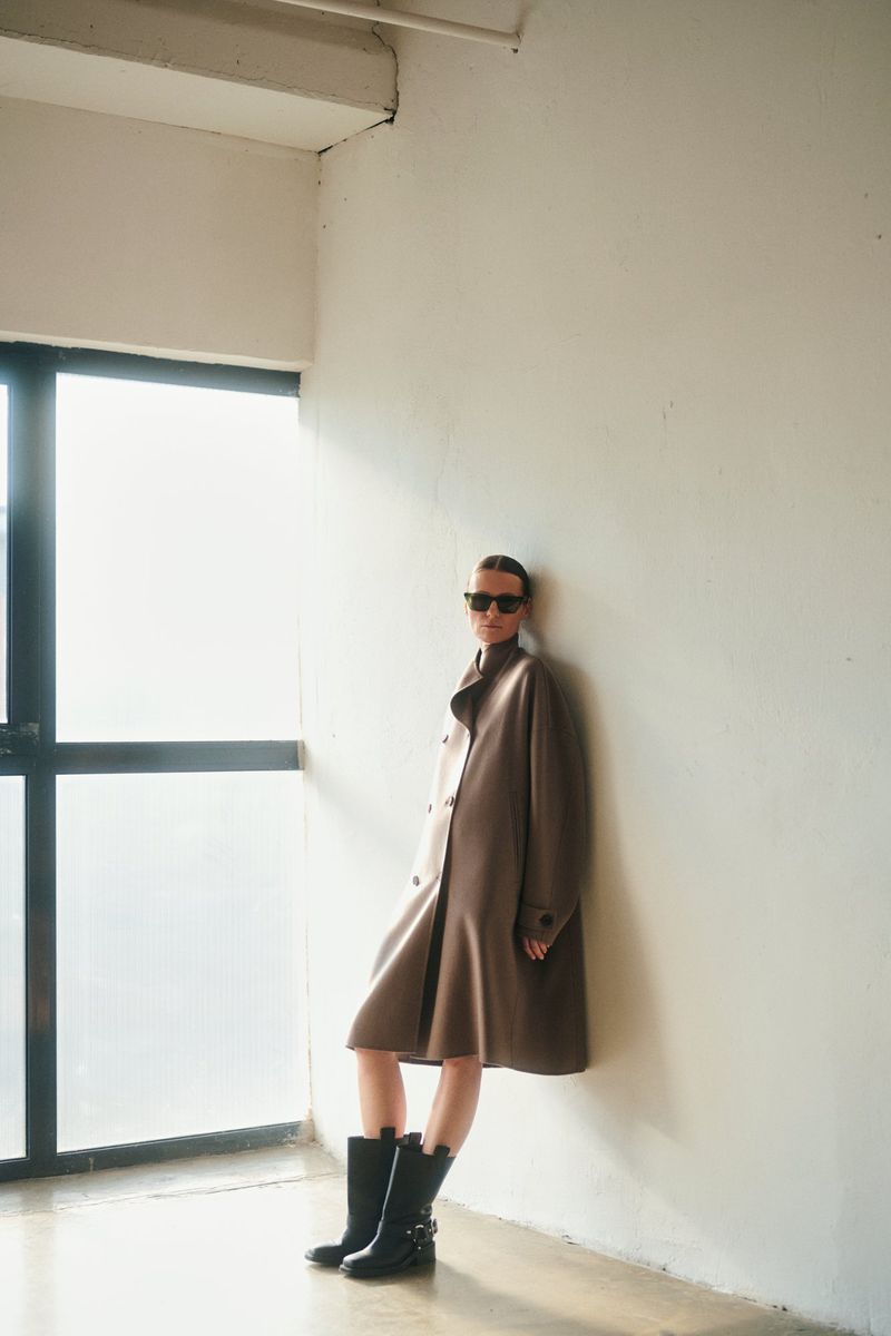 Model wearing dark sunglasses, a brown coat, and black boots leaning against a plain wall near a large window.