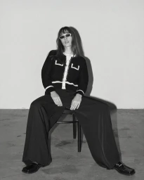 Model seated on a chair wearing a dark cardigan and wide-leg trousers against a plain studio background.
