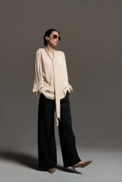Model wearing a cream blouse and black wide-leg trousers against a neutral grey studio backdrop.