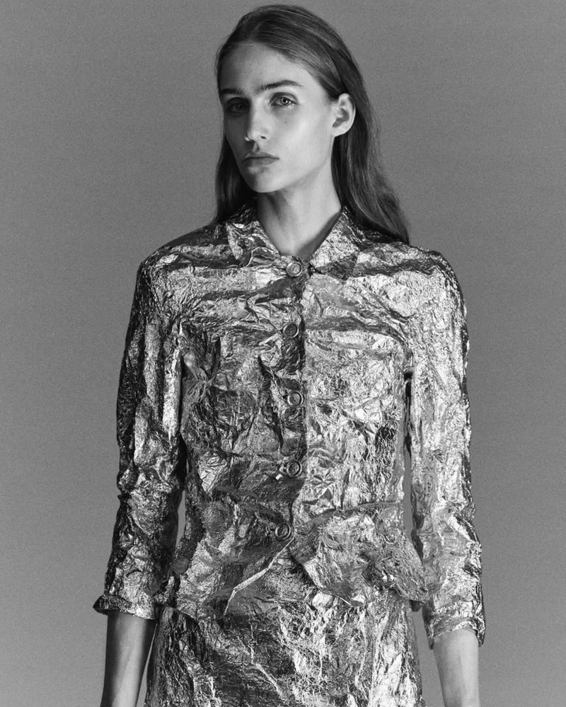 Monochrome portrait of a young woman in a reflective silver jacket and skirt, posing in a photo studio with soft lighting and a neutral backdrop.