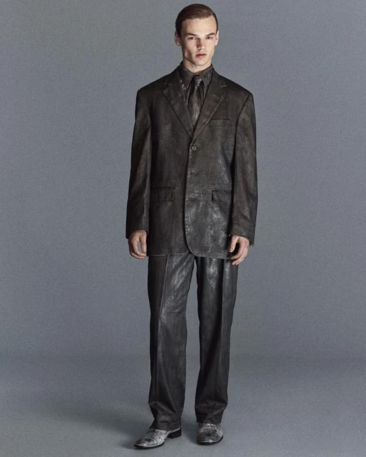 Adult male in a dark-grey suit and tie stands in front of a plain grey background in a fashion photography studio setting.