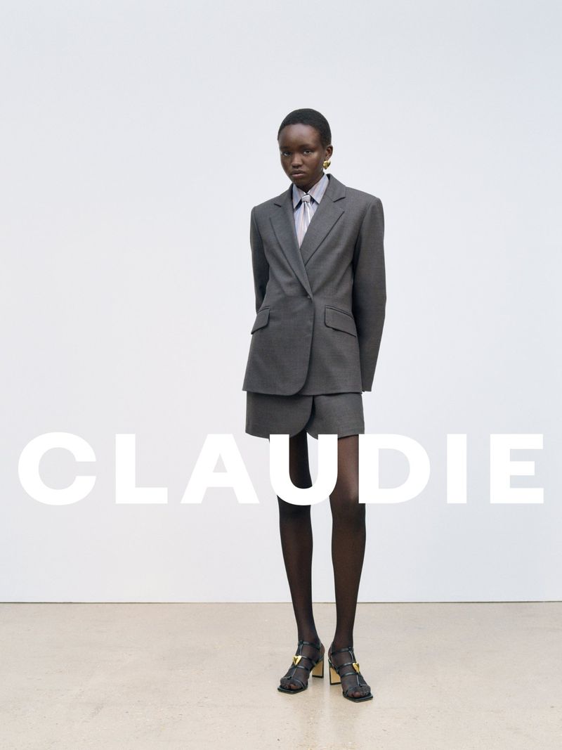 A tall, thin African woman posing indoors wearing a gray suit with shorts and heels against a white wall with the brand name CLAUDIE.