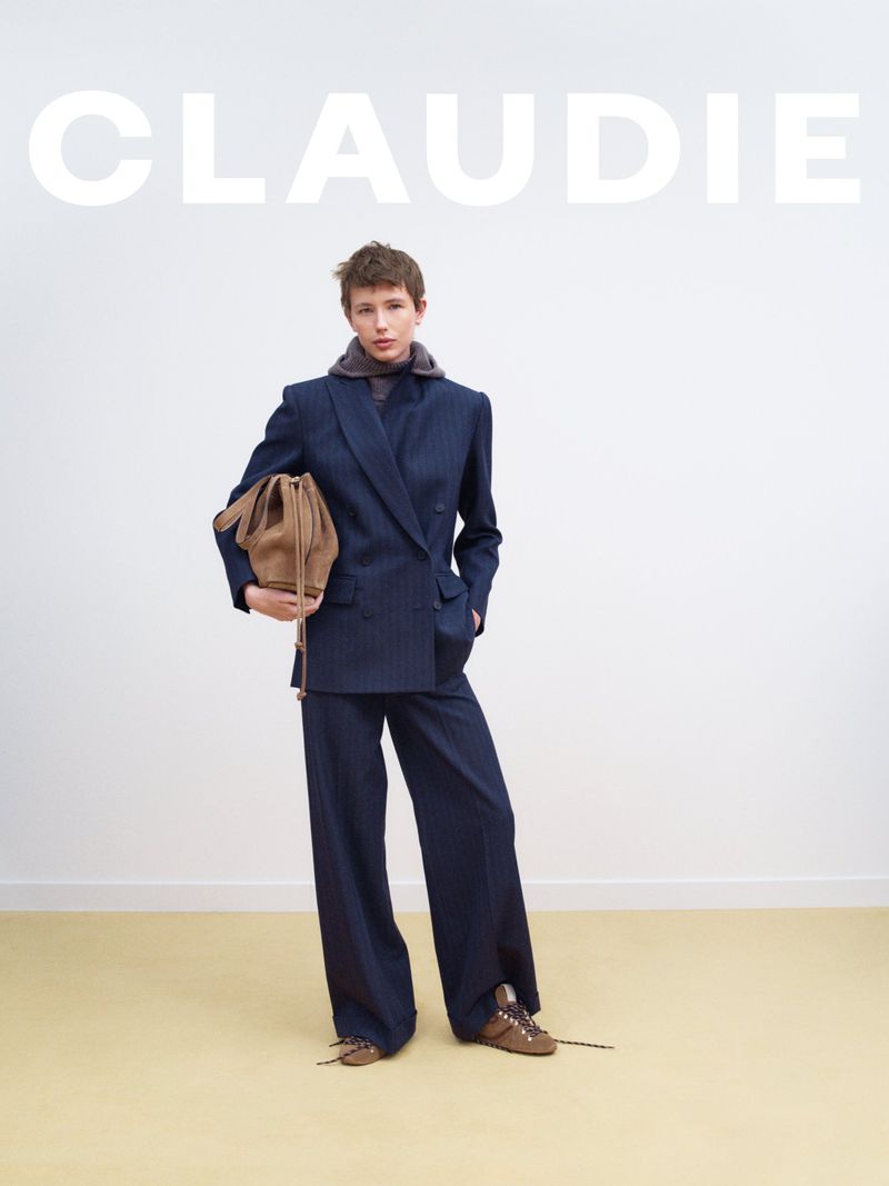 An adult woman poses indoors against a white wall wearing a navy suit, brown sneakers and holding a tan-colored drawstring bag.