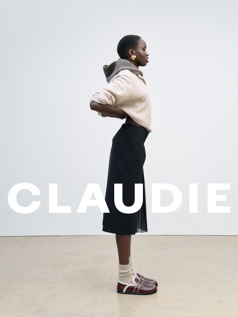 A full-length studio shot of a Black woman wearing a beige hooded sweater, black skirt, socks, and maroon shoes in a fashion editorial.