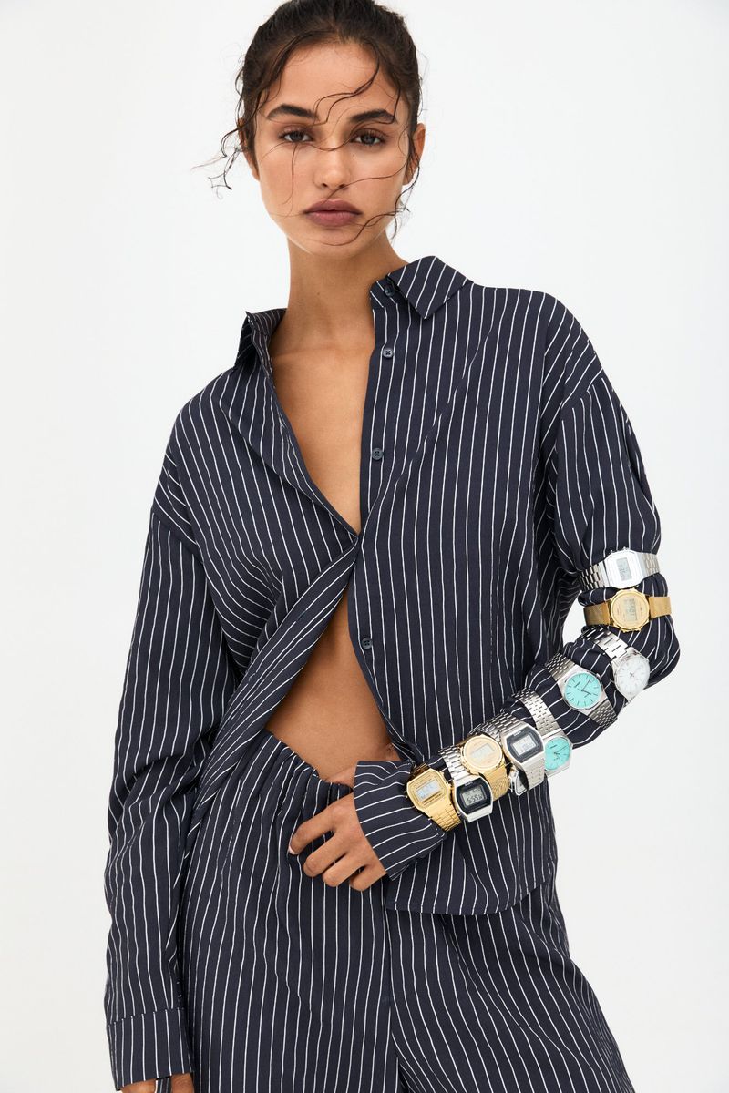 Fashion shot of a woman in a striped shirt and pants, with multiple watches on her arm, against a white backdrop in soft studio lighting.
