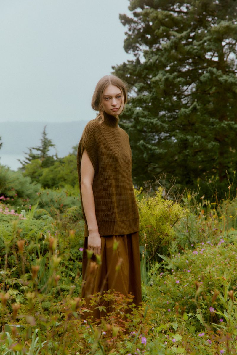 An adult woman models olive green sleeveless sweater and skirt in a grassy outdoor setting with trees and overcast lighting.