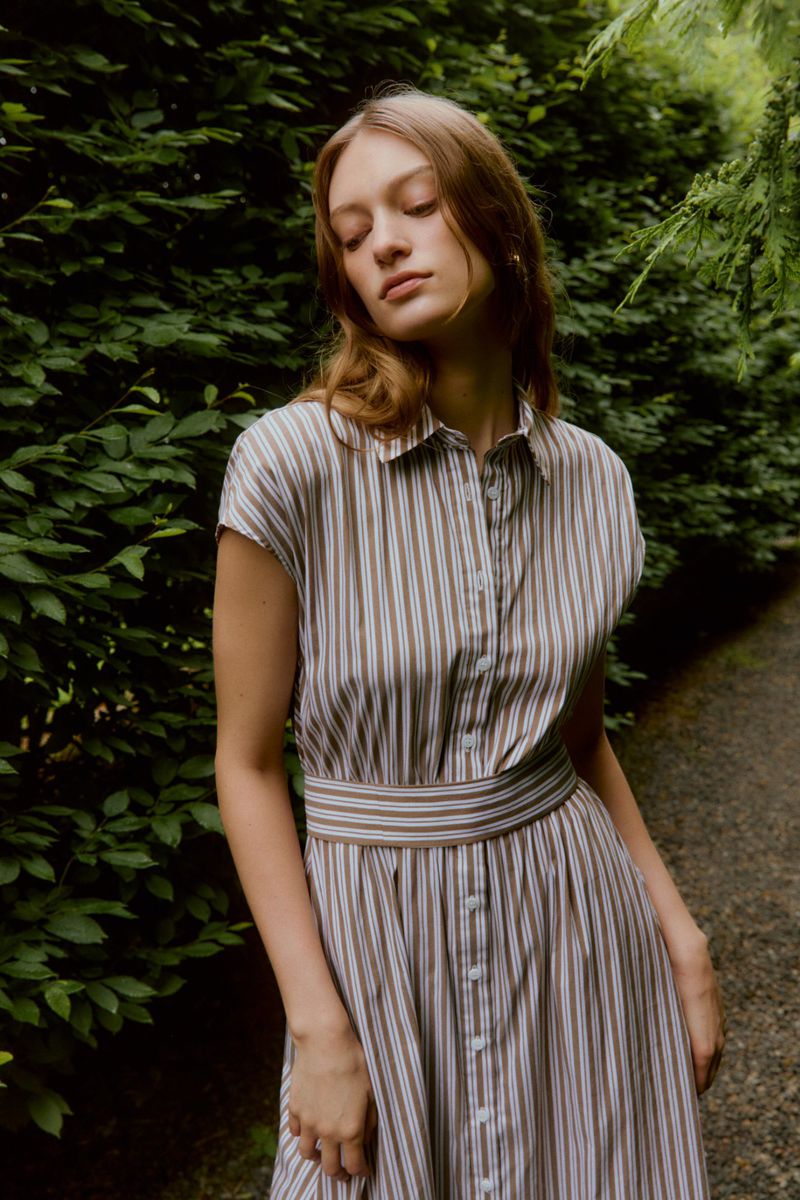 A woman with light brown hair poses in a striped shirt dress outdoors with a leafy green background, creating a natural and soft fashion image.