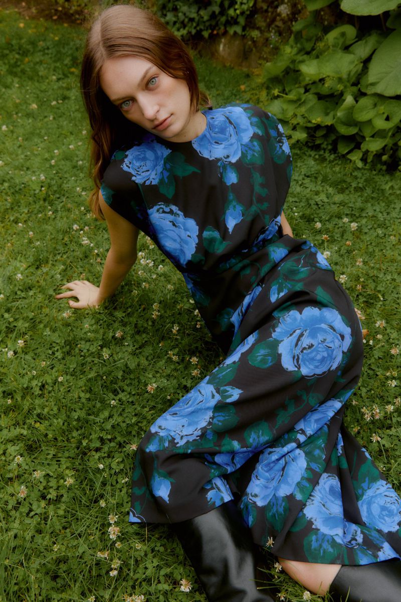 Young woman in a floral dress with black boots sitting on a lawn in a natural setting, gazing at the camera with a neutral expression.