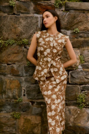 Adult woman posing outdoors in brown floral-print skirt suit, standing against a stone wall with greenery, looking to the side.