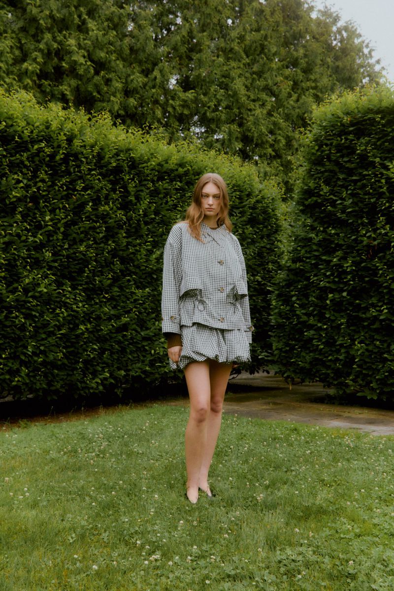 An adult female stands on a lawn surrounded by green foliage. She is wearing a matching checkered jacket and skirt. Outdoor setting.