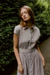 Model Cleirys Velasquez wearing a brown and white striped belted shirt dress outdoors against lush green garden hedges.