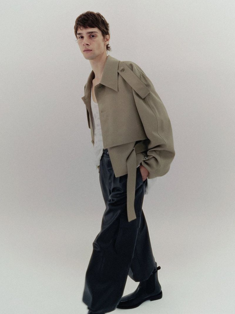 Model Patryk Lawry wearing a cropped beige belted jacket, wide black trousers, and black boots against a white studio background.