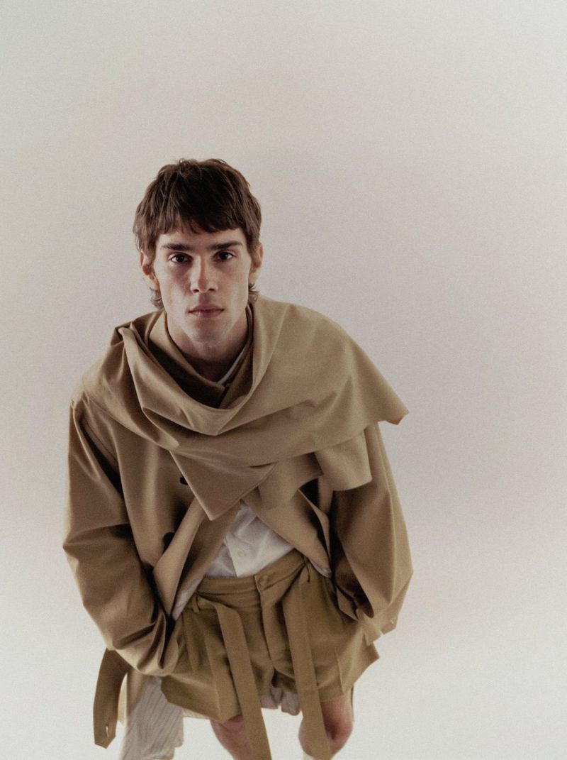 A young adult male is posing in a studio with a tan overcoat, shorts, and a neutral background, creating a minimalist fashion image.