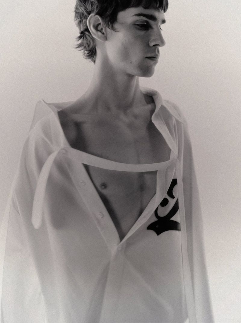 Monochrome portrait of a slender adult male wearing an open white shirt with a distinctive black graphic, looking away from the camera.