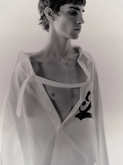 Monochrome portrait of a slender adult male wearing an open white shirt with a distinctive black graphic, looking away from the camera.