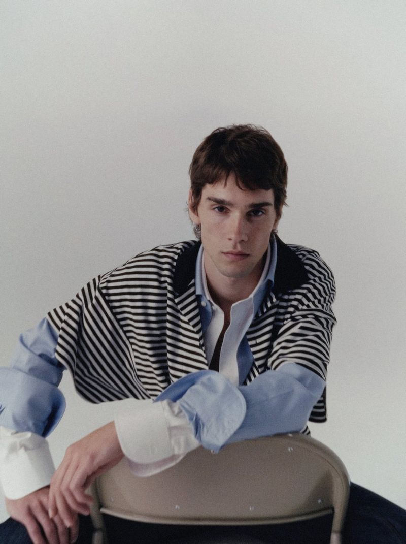 Portrait of a young adult man in a striped shirt and light blue button-down, sitting on a metal chair against a neutral backdrop.