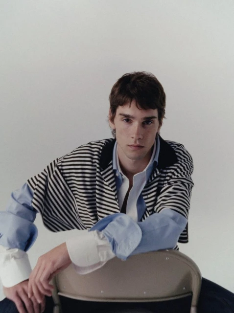 Portrait of a young adult man in a striped shirt and light blue button-down, sitting on a metal chair against a neutral backdrop.