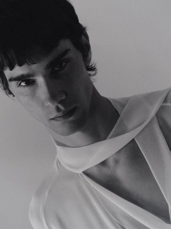 Close-up portrait of a male model wearing a white top with a draped asymmetric neckline against a plain neutral background.
