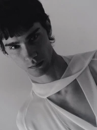Close-up portrait of a male model wearing a white top with a draped asymmetric neckline against a plain neutral background.