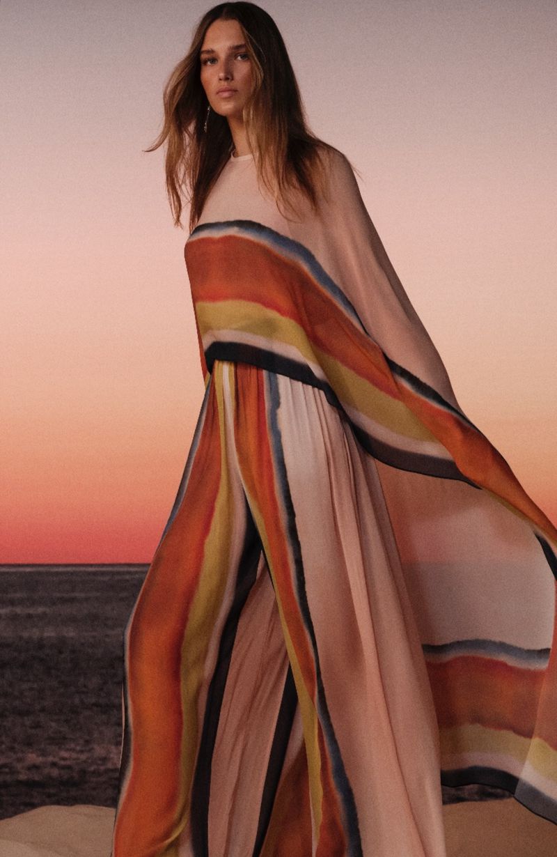 Woman in flowing striped dress poses on sand dune against sunset. Fashion shot with desert and sky backdrop for a clothing brand advertisement.