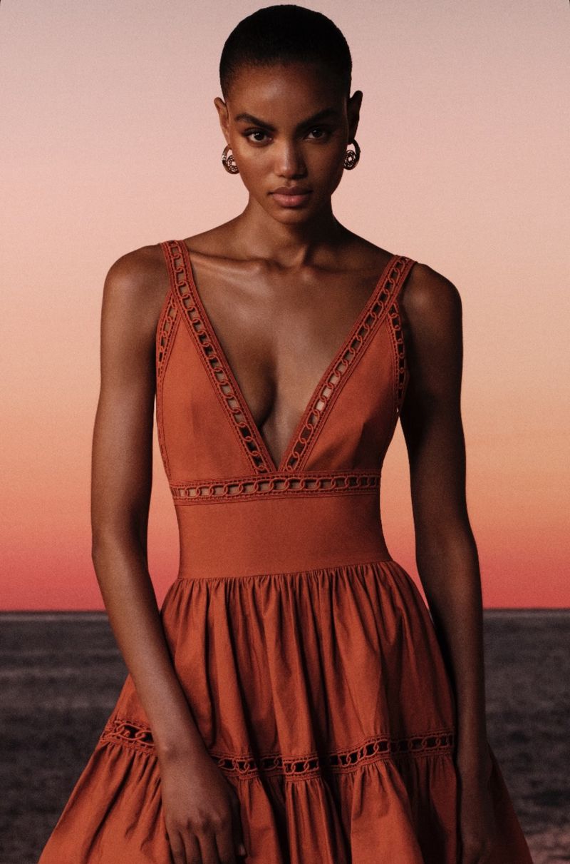 A fashionable woman with short hair poses outdoors in a sleeveless, burnt-orange dress with a V-neck and decorative cutouts.