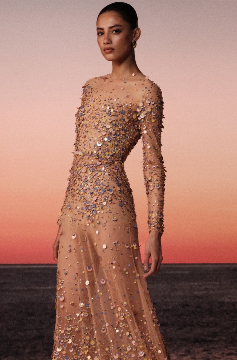 Elegant woman in sequined gown stands against a gradient peach sky and dark shoreline. The long-sleeved dress is fitted and detailed.
