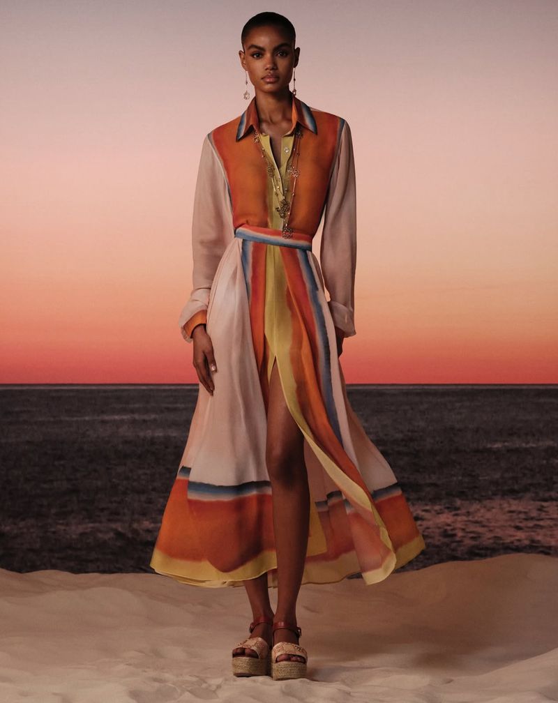 Adult female fashion model posing in long, colorful shirt dress on beach at dusk with a sea and pink sunset in the background.