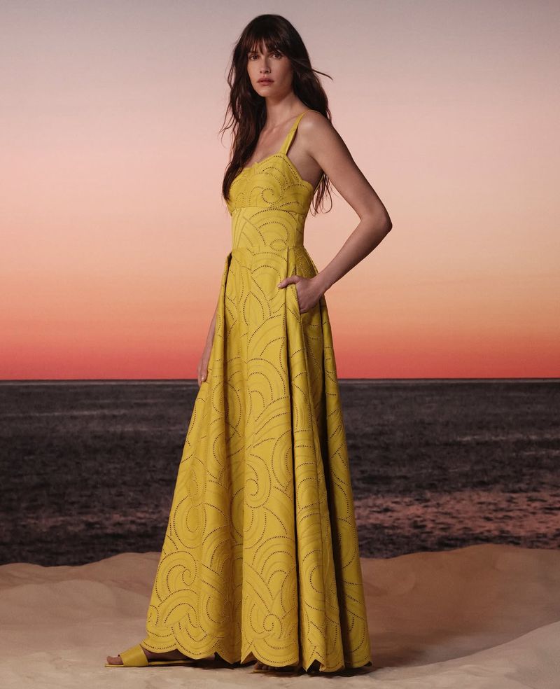A brunette woman wearing a long yellow dress with an ocean sunset background. The dress has a pocket and subtle wave pattern.