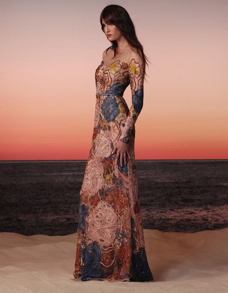 A brunette woman in a long, patterned gown stands in a beach setting with a pink and orange sunset sky in the background.
