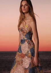 Model wearing a strapless floral embroidered gown standing against a sunset sky and ocean horizon in a lookbook image