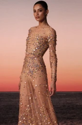 Model wearing a sheer long-sleeve gown covered in shimmering multicolored sequins against a sunset sky and dark horizon