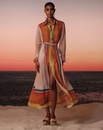 Model with buzzcut hair posing on a sand dune in a flowing sunset-toned shirt dress against an ocean horizon at dusk.