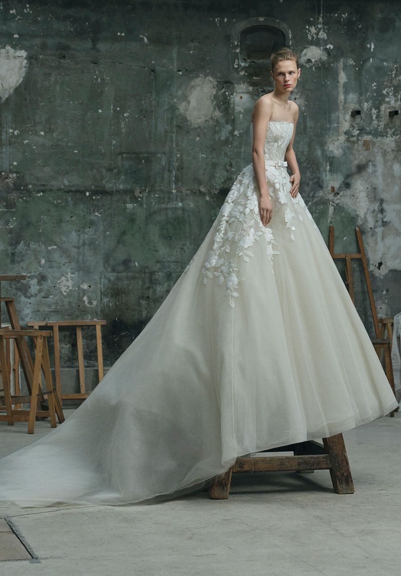 Woman poses in a white strapless ballgown with floral embroidery in a studio with a distressed wall and wooden props.