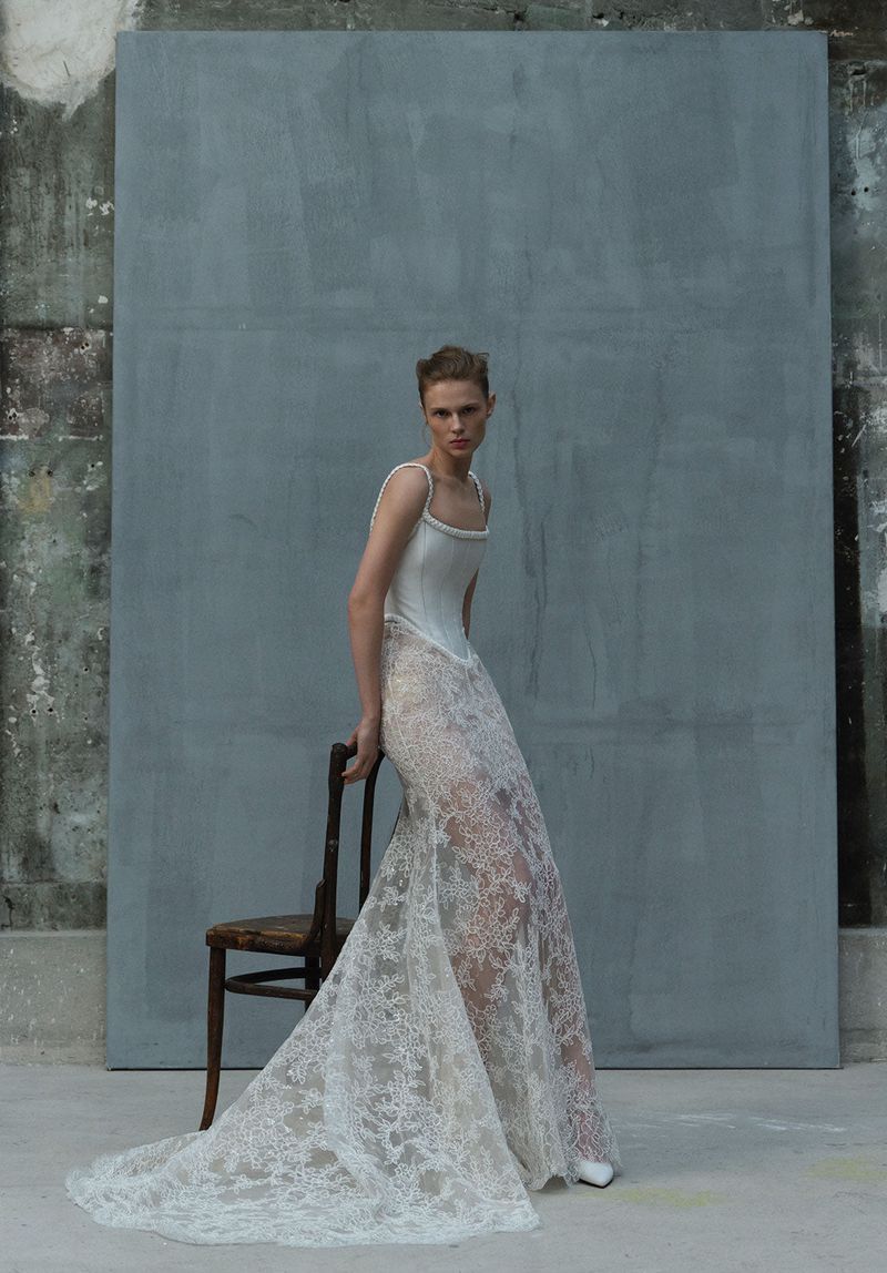 A model in a flowing lace gown leans against a wooden chair against a mottled gray backdrop in a fashion editorial setting.