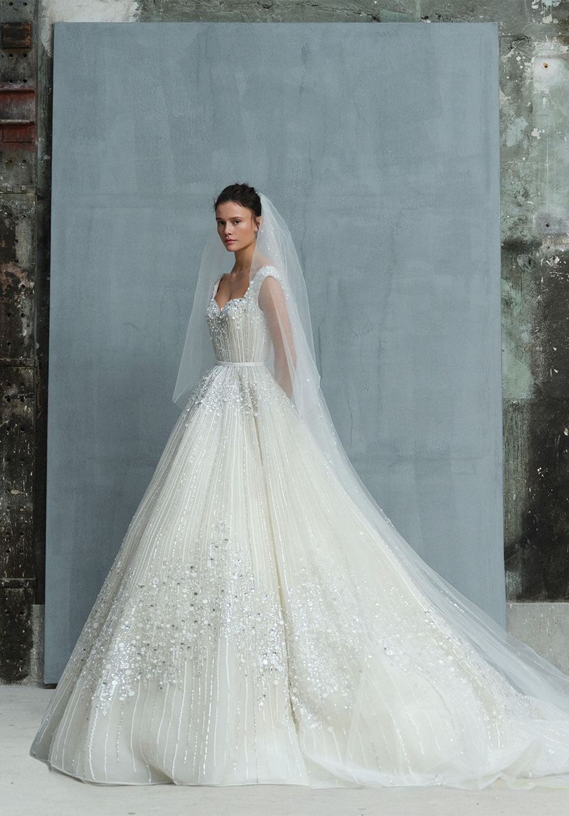 Woman in ornate white wedding dress with veil posing in a bright indoor photo studio with neutral background.