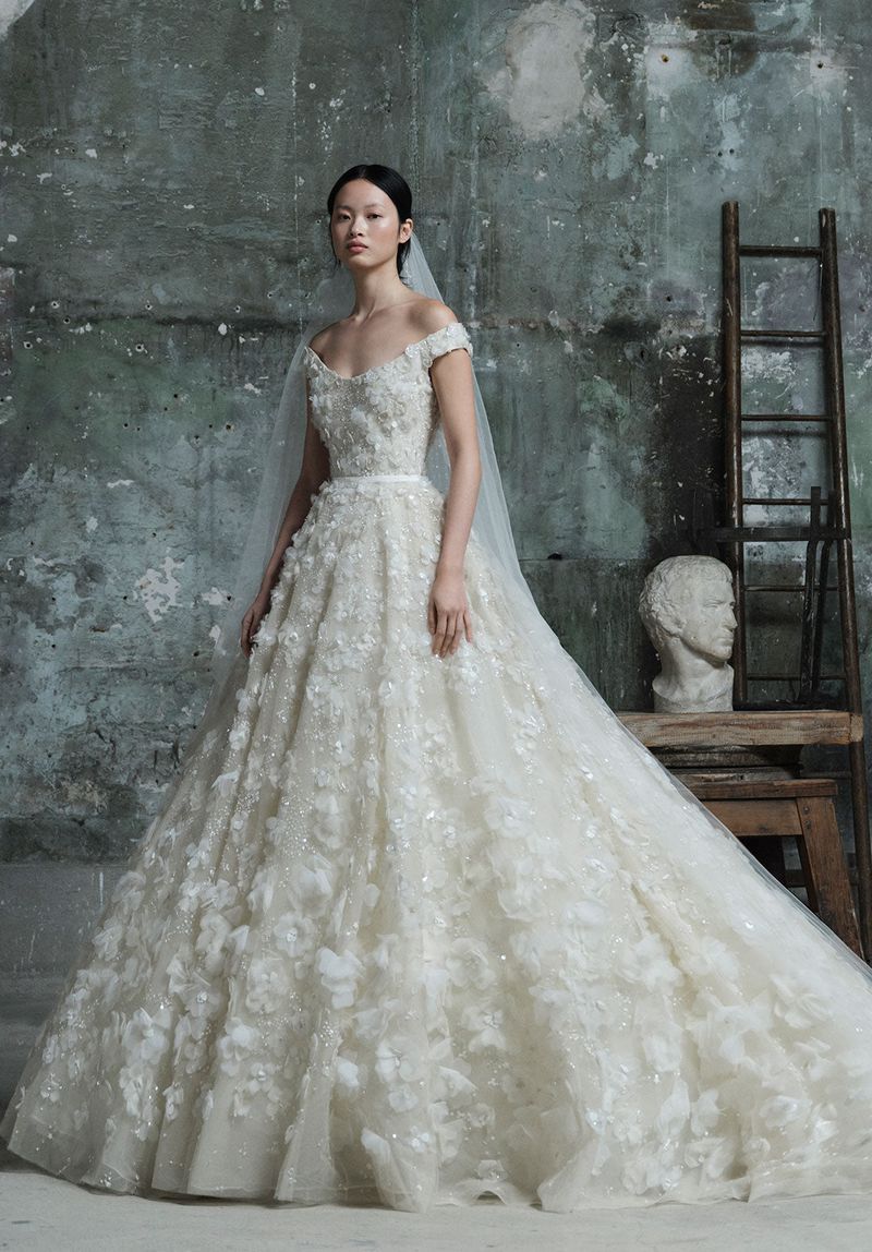 Elegant woman in a white floral wedding gown and veil standing in front of a textured wall, captured in soft, diffused lighting.