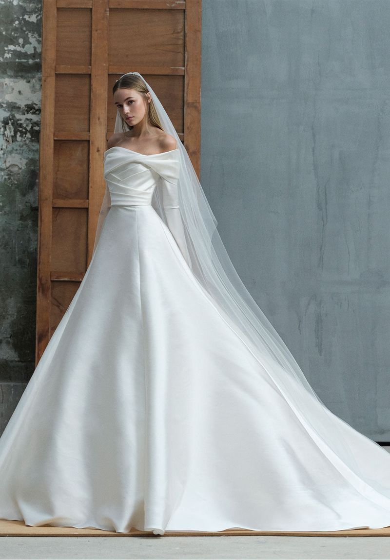 A woman wears a white off-the-shoulder wedding gown and veil, posing against a neutral backdrop, illuminated by soft studio lighting.