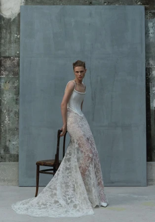 Model posing in an Elie Saab white lace bridal gown with corset top next to a wooden chair against a grey panel backdrop.